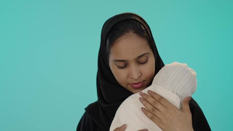 أجواء عائلية مليئة بالحب والحنان، الرعاية والاهتمام بالابناء، بورتريه مقرب لسيدة عربية خليجية سعودية سمراء ترتدي الحجاب و العباءة السوداء تحمل طفلتها الرضيعة بحب، خلفية خضراء