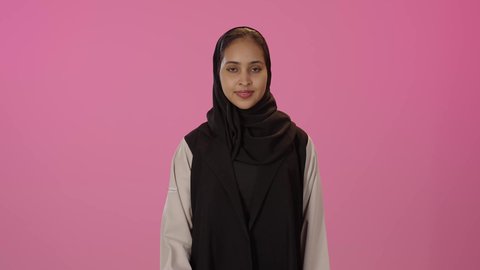 The concept of acceptance and admiration, looking at the camera with expressions of joy, a sign of quality and excellence, a portrait of a young Arab Gulf Saudi woman with a dark complexion, smiling, wearing a hijab and casual attire, raising her thumb up, with a colorful background.