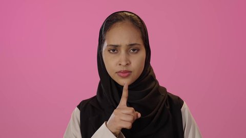مفهوم التوقف عن الكلام وخفض الصوت، الهدوء والسكون وعدم الإزعاج، بورتريه مقرب لشابة عربية خليجية سعودية محجبة سمراء ترتدي الزي الكاجوال تضع أصبع السبابة على فمها بإيماءات طلب السكوت والصمت، خلفية ملونه