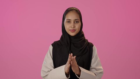 الاهتمام بالمظهر الخارجي والأناقة والأنوثة، مفهوم الاستقبال والترحيب، الإكثار من الدعاء في شهر رمضان، بورتريه مقرب لامرأة عربية خليجية سعودية سمراء مبتسمه ترتدي الحجاب والزي الكاجوال تنظر إلى الكاميرا ترفع يديها بايماءات الدعاء والتضرع, خلفية ملونه