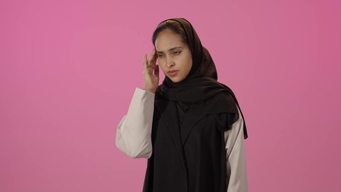 الشعور بألم الرأس والصداع، الحاجة إلى العلاج والمساعده، المعاناة من آلام الشقيقة، الحاجة للهدوء والراحة، بورتريه لشابة خليجية سعودية سمراء محجبة ترتدي الزي الكاجوال تضع يديها على رأسها بايماءات التعب والارهاق، خلفية ملونه