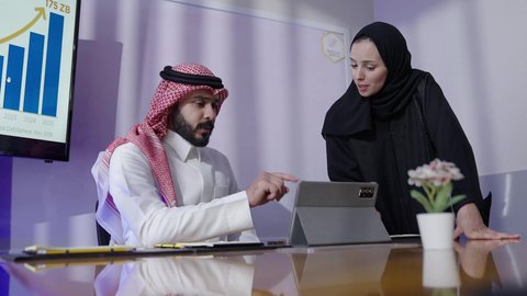 دمج التقنيات المتطوره فى تطوير الأعمال وحل المشكلات، مراجعة الخطط والاستراتيجيات والاتفاقيات عبر التقنيات الحديثه، مكاتب و شركات خليجية، رجل عربي سعودي خليجي يرتدي الثوب التقليدي والشماغ  مع موظفه سعوديه يجلسون في المكتب داخل مقر الشركة يستخدمون جهاز التابلت ،مهنة ووظيفة سعوديه