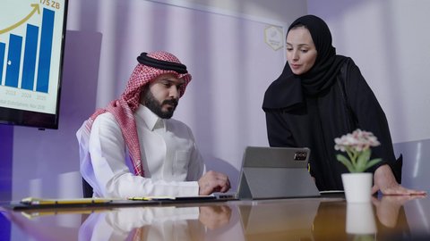 Reviewing plans, strategies, and agreements using modern technologies. Gulf offices and companies. A Saudi Arabian man wearing a traditional thobe and shemagh sits with a Saudi female employee in an office at the company headquarters using a tablet. Saudi profession and job. Unity and teamwork for business development and problem solving.