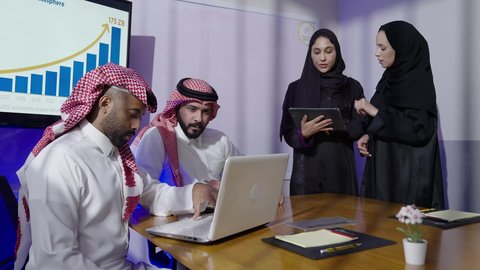 Reviewing plans, strategies, and agreements. Saudi profession and job. Unity and teamwork for business development and problem solving. Gulf offices and companies. Two Saudi Arabian Gulf men wearing traditional thobes and shemaghs with two Saudi employees sitting in an office inside the company headquarters using a laptop and tablet.