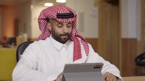 دمج التقنية والتكنولوجيا في الحياة العملية، استخدام التقنيات الحديثة المتطورة، دراسه المشروع ومراجعه التقرير السنوي،جلسه عمل عن بعد, أداء المهمة اليومية داخل مقر الشركه، رجل أعمال عربي خليجي سعودي يرتدي الثوب التقليدي والشماغ يقوم بالعمل على جهاز التابلت