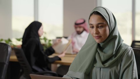 دراسه المشروع ومراجعه التقرير السنوي، استخدام التقنيات الحديثة المتطورة، جلسه عمل عن بعد, أداء المهمة اليومية داخل مقر الشركه، امراة عربية خليجية سعودية محجبة ترتدي العباءة الملونة تقوم بالعمل على جهاز الحاسوب المكتبي، دمج التقنية والتكنولوجيا في الحياة العملية