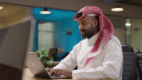رجل أعمال عربي خليجي سعودي يرتدي الثوب التقليدي والشماغ يقوم بالعمل على جهاز الحاسوب المحمول داخل مقر الشركه، دمج التقنية والتكنولوجيا في الحياة العملية، أداء المهمة اليومية داخل مقر الشركه، دراسه المشروع ومراجعه التقرير السنوي، جلسه عمل عن بعد