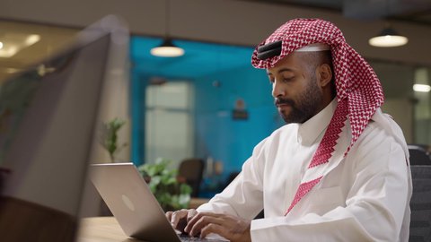 Integrating technology into practical life, performing daily tasks at company headquarters, studying a project and reviewing an annual report, a remote work session, a Saudi Arabian Gulf businessman wearing a traditional thobe and shemagh working on a laptop at company headquarters.