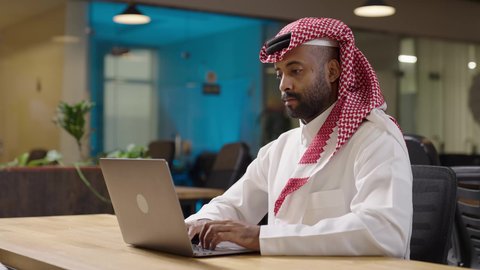 Performing daily tasks at company headquarters, studying the project and reviewing the annual report, remote working session, a Saudi Arabian Gulf businessman wearing a traditional thobe and shemagh working on a laptop at company headquarters, integrating technology into work life.