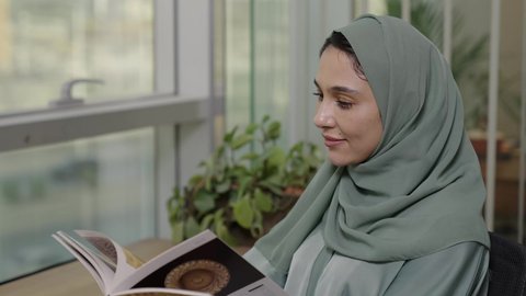 ممارسة هواية المطالعة, مفهوم الشغف بالقراءة, امراة عربية خليجية سعودية محجبة ترتدي العباءة الملونة تجلس على الطاولة و تمسك كتاب بيديها, الكتب و الروايات على المسلية, أماكن هادئة مريحة للدراسة