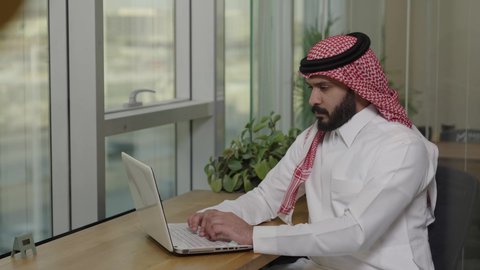 دراسه المشروع ومراجعه التقرير السنوي، استخدام التقنيات الحديثة المتطورة، جلسه عمل عن بعد, أداء المهمة اليومية داخل مقر الشركه، رجل أعمال عربي خليجي سعودي يرتدي الثوب التقليدي والشماغ يقوم بالعمل على جهاز الحاسوب المحمول داخل مقر الشركه، دمج التقنية والتكنولوجيا في الحياة العملية
