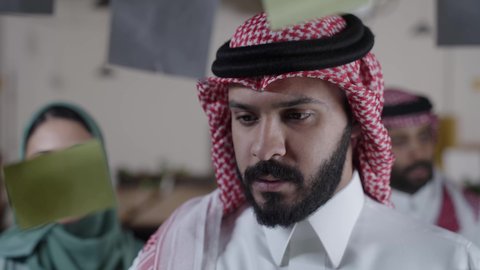 Writing notes and tasks on a sticky note, colored paper stickers on glass, modern meeting room, Saudi Gulf Arab employees inside the company headquarters, productive work environment, Saudi office and company, participating in goal setting, Saudi professions and jobs