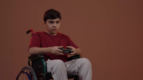 إيماءات الاستمتاع والاندماج، استخدام جهاز تكنولوجي تقني حديث ومتطور، قضاء وقت ممتع في ألعاب الفيديو الترفيهيه، بورتريه لولد عربي خليجي سعودي يرتدي الزي الكاجوال يجلس على كرسي متحرك يحمل بيده عصا التحكم