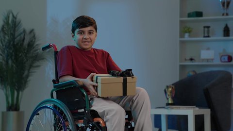 مفهوم الموده والالفه ، الشعور بالسعادة والفرح, بورتريه لولد عربي خليجي سعودي مبتسم يرتدي الزي الكاجوال يجلس على كرسي متحرك ينظر إلى الكاميرا يحمل بيديه علبة هدية مغلفة، مناسبات وأعياد سعيدة، تقديم الهدايا والتهنئات