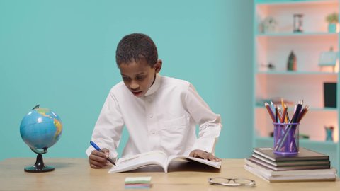 Back to school, reviewing lessons diligently and diligently to achieve success and excellence, focusing on solving and writing assignments, a Saudi Arabian Gulf student wearing traditional clothing sitting at a desk