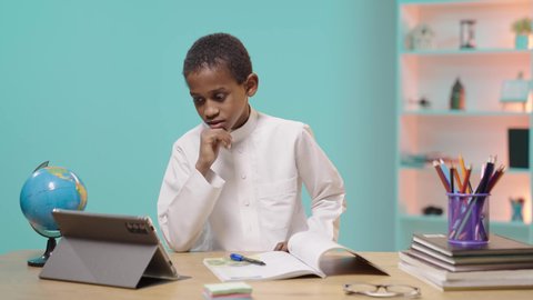 Using modern technical devices, following classes and solving assignments online, studying and studying remotely, a Saudi Arabian Gulf student wearing traditional clothing sitting at a desk and reviewing lessons using a tablet