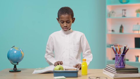 A Saudi Arabian Gulf student wearing traditional clothing sits at a desk holding a lunch box and starts eating. Meals rich in important nutrients for building the body, back to school season.