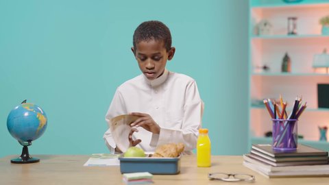 الوجبات الغنية بالعناصر الغذائية الهامة لبناء الجسم،  موسم العودة إلى المدارس،  طالب عربي خليجي سعودي  يرتدي الزي التقليدي يجلس على مكتب يقوم بامساك لانش بوكس ويبدأ بالاكل