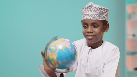 مفهوم البحث والإستكشاف حول العالم، الإستعداد للعام الدراسي الجديد، العودة إلى المدارس, صبي عربي خليجي عماني مبتسم يرتدي الدشداشة والكمة يقوم بالامساك بمجسم الكرة الارضية, خلفية زرقاء