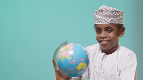 خريطة العالم الكروية, مفهوم البحث والاستكشاف حول العالم, العودة إلى المدارس, بورتريه لصبي عربي خليجي عماني مبتسم يرتدي الدشداشة والكمة يقوم بالامساك بمجسم الكرة الارضية, خلفية زرقاء