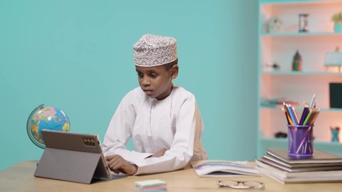 تطور العلم والتعليم في سلطنه عمان، بورتريه لتلميذ عربي خليجي عماني يرتدي الدشداشه والكمه يجلس أمام طاولة المذاكرة و يقوم بالنقر على جهاز التابلت, استخدام الأجهزة والتقنيات الحديثة والمتطورة, مفهوم التعليم عن بعد, خلفية زرقاء