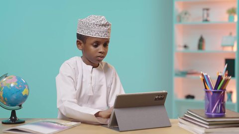 مراجعة الدروس اليومية عبر الانترنت، تطور العلم والتعليم في سلطنه عمان، بورتريه لتلميذ عربي خليجي عماني يرتدي الدشداشه والكمه يجلس أمام طاولة المذاكرة و يقوم بتدوين المعلومات على جهاز التابلت, خلفية زرقاء