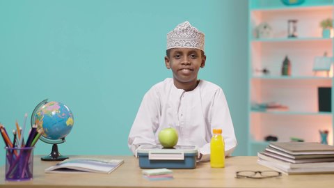 موسم العودة للمدارس، الاستمتاع بتناول الأطعمة الصحية في وقت الفسحة، الوجبات الغنية بالعناصر الغذائية الهامة لبناء الجسم، تلميذ عربي خليجي عماني مبتسم يرتدي الدشداشه والكمه يجلس في مقعده امامه اللانش البوكس, خلفية زرقاء