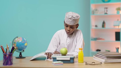 الاستمتاع بتناول الأطعمة الصحية في وقت الفسحة، الوجبات الغنية بالعناصر الغذائية الهامة لبناء الجسم، تلميذ عربي خليجي عماني مبتسم يرتدي الدشداشه والكمة يجلس في مقعده امامه تفاحة خضراء و اللانش بوكس, خلفية زرقاء