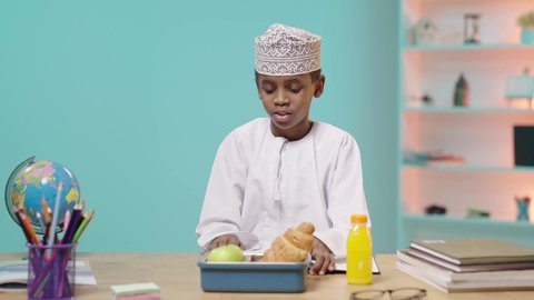 موسم العودة للمدارس، الاستمتاع بالأطعمة في وقت الفسحة، تناول قطعه من المخبوزات, تلميذ عربي خليجي عماني يرتدي الدشداشه والكمه يجلس في مقعده امامه اللانش البوكس, خلفية زرقاء