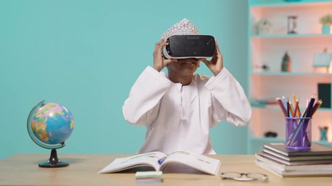 دمج الواقع الافتراضي بالتعلم والتدريب، استخدام التقنيات الحديثة في التعليم المدرسي, ولد عربي خليجي عماني يرتدي الدشداشة و الكمة يجلس في مقعده و يضع نظارة الفي ار ثلاثية الأبعاد, خلفية زرقاء