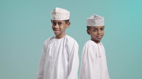 الوقوف باستقامة بإيماءات أيدي متقاطعة، بورتريه لولدين عربيين خليجيين عمانيين يرتديان الدشداشة و الكمة ينظران الى الكاميرا بتعابير الفرح والسرور, قضاء وقت ممتع برفقة الأصدقاء, خلفية زرقاء