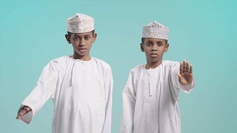 الشعور بالإنزعاج والغضب، بورتريه لولدين عربيين خليجيين عمانيين يرتديان الدشداشة و الكمة يقومان برفع يدهم بإيماءات التوقف وعدم الاقتراب, التعبير عن موقف سلبي تجاه شيئاً ما, خلفية زرقاء