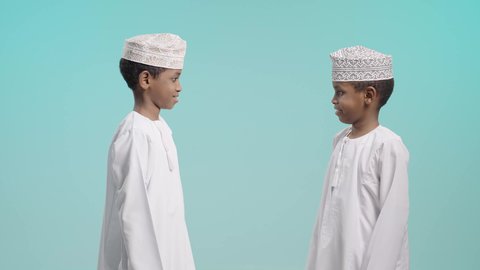 لحظة مرح و تفاعل بين الأصدقاء, الصداقة والبهجة و روح التعاون, بورتريه لولدين عربيين خليجيين عمانيين يرتديان الدشداشة و الكمة يتبادلان ضربة يد مليئة بالحيوية, خلفية زرقاء