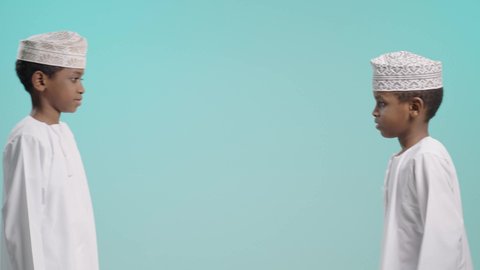 ابتسامة وعناق يرمزان للوفاء، بورتريه لولدين عربيين خليجيين عمانيين يرتديان الدشداشة و الكمة يقوم أحدهما باحتضان الاخر, لحظة دفء تعبّر عن الألفة والمودة, خلفية زرقاء