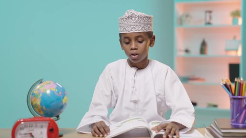 الثقافة والقراءة والإستطلاع، مراجعة الدروس اليومية، طالب عربى خليجي عماني يرتدي الدشداشة و الكمة يجلس على طاولة مكتبية يقوم بالمذاكرة, ايماءات التركيز والاندماج, خلفية زرقاء