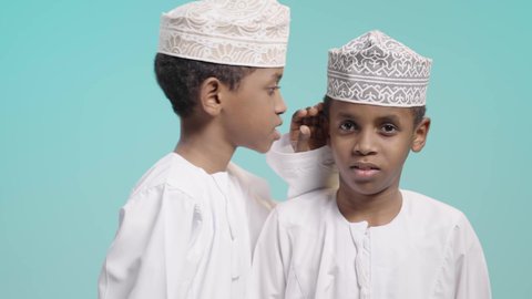 Speaking in a low voice, facial expressions indicating integration and happiness, a portrait of two Omani Gulf Arab boys wearing a dishdasha and a kummah, one of them whispering in his brother's ear, keeping and preserving secrets, blue background