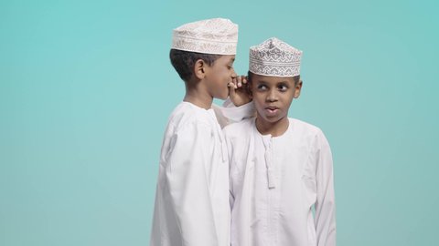 ايماءات وجه تدل على الاندماج والسعادة, بورتريه لولدين عربيين خليجيين عمانيين يرتديان الدشداشة و الكمة يقوم أحدهما بالهمس في اذن اخوه, كتمان وحفظ الاسرار, التحدث بصوت منخفض, خلفية زرقاء