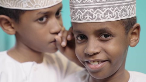 كتمان وحفظ الاسرار، بورتريه لولدين عربيين خليجيين عمانيين يرتديان الدشداشة و الكمة يقوم أحدهما بالهمس في اذن اخوه، التحدث بصوت منخفض، ايماءات وجه تدل على الاندماج والسعادة, خلفية زرقاء