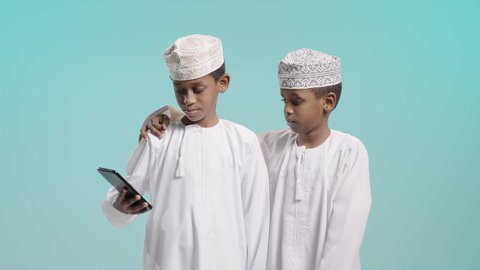 قضاء وقت ممتع برفقة الأصدقاء، صديق يضع يده على كتف صديقه, بورتريه لولدين عربيين خليجيين عمانيين يرتديان الدشداشة و الكمة يقومان بتصفح شئ على شاشة الهاتف المحمول, خلفية زرقاء