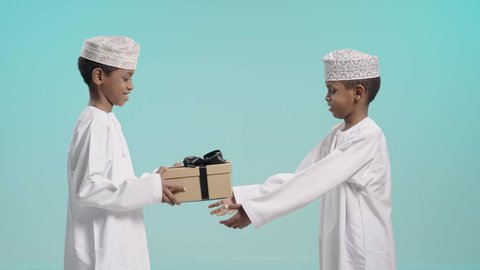 مشاركة وتقديم الهدايا في الأعياد والمناسبات السعيدة، التعبير عن الحب والتقدير والاحترام، بورتريه لولدين عربيين خليجيين عمانيين يرتديان الدشداشة و الكمة يقوم أحدهما بإهداء هدية للاخر, خلفية زرقاء