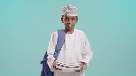 Looking at the camera with gestures of happiness and joy, seriousness and diligence to achieve success, the concept of returning to school, a portrait of an Arab Gulf Omani student wearing a dishdasha and a kummah carrying a school backpack and holding books in his hand, blue background