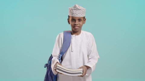 الجد والاجتهاد للوصول إلى النجاح، مفهوم العودة إلى المدارس، بورتريه لطالب عربي خليجي عماني يرتدي الدشداشة والكمة يحمل حقيبة الظهر المدرسية يمسك بيده كتب, النظر الى الكاميرا بإيماءات السعادة والفرح, خلفية زرقاء