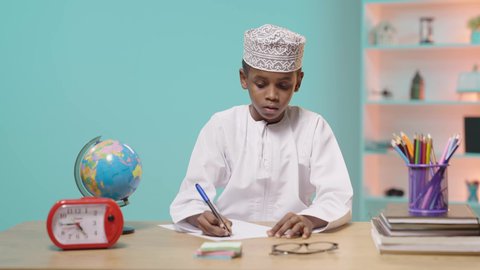 The concept of diligence and striving for excellence, modern and developed educational curricula and courses, the back-to-school season,  an Arab Gulf Omani student wearing a dishdasha and a kummah sitting at an office table studying his school lessons, blue background