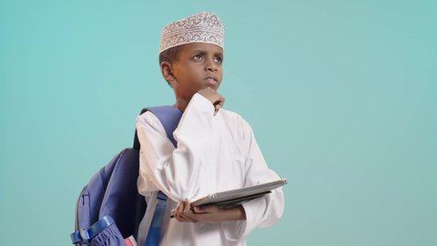 Studying and studying remotely, using modern technical devices, following classes and solving assignments online, a portrait of an Arab Gulf Omani student wearing a dishdasha, a turban, and a school bag, reviewing lessons using a tablet, blue background