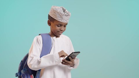 متابعة الحصص عبر الأونلاين, مفهوم التعليم عبر الانترنت, استخدام الأجهزة والتقنيات التكنولوجية, بورتريه لولد عربي خليجي عماني يرتدي الدشداشة والكمة وحقيبة الظهر المدرسية يمسك الهاتف المحمول بيده بإيماءات التركيز والاندماج, خلفية زرقاء
