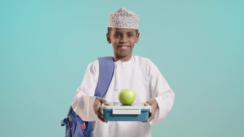 الوجبات الغنية بالعناصر الغذائية الهامة لبناء الجسم، موسم العودة إلى المدارس، بورتريه لطالب عربي خليجي عماني مبتسم يرتدي الدشداشة والكمة والحقيبة المدرسية  يقوم بامساك لانش بوكس وتفاح, خلفية زرقاء