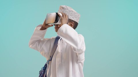 The concept of simulating the imaginary world, integrating virtual reality with learning and training, using modern technologies in school education, a portrait of an Arab Gulf Omani boy wearing a dishdasha, a turban, and a backpack, wearing 3D VR glasses, blue background
