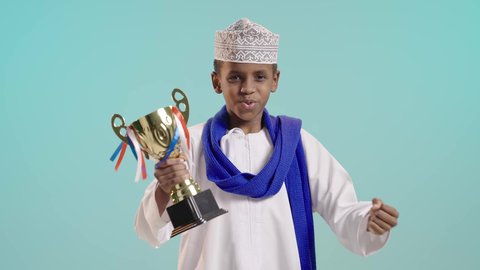 تشجيع الفريق العماني، الفرح والسرور لفوز المنتخب الوطني، ايماءات الحماس والتشجيع,  بورتريه لولد عربي خليجي عماني يرتدي الدشداشة والكمة ووشاح أزرق يمسك بيده كأس الفوز و تظهر عليه تظهر عليه تعابير السعادة بالانتصار, خلفية زرقاء