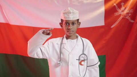 تحديد واختيار مهنة المستقبل منذ الصغر، الطموح والهدف المستقبلي في العمل في القطاع الطبي في المستشفى، ولد عربي خليجي عماني يرتدي الكمة والمعطف الأبيض والسماعة الطبية يرفع يده لالقاء التحية, الحلم المستقبلي ليكون طبيب ناجح, خلفية علم سلطنة عمان
