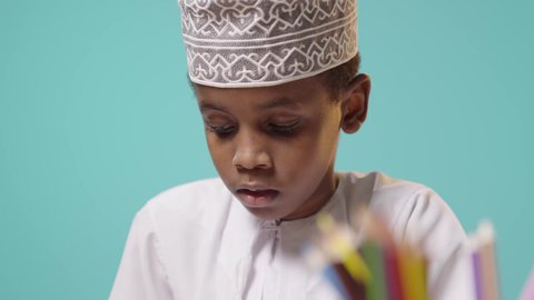 ايماءات الاندماج والتركيز, استخدام أقلام التلوين الخشبية, بورتريه لولد صغير عربي خليجي عماني يرتدي الدشداشة والكمة يقوم بالكتابة والتلوين, استغلال وقت الفراغ في التعليم والتدريب, خلفية زرقاء